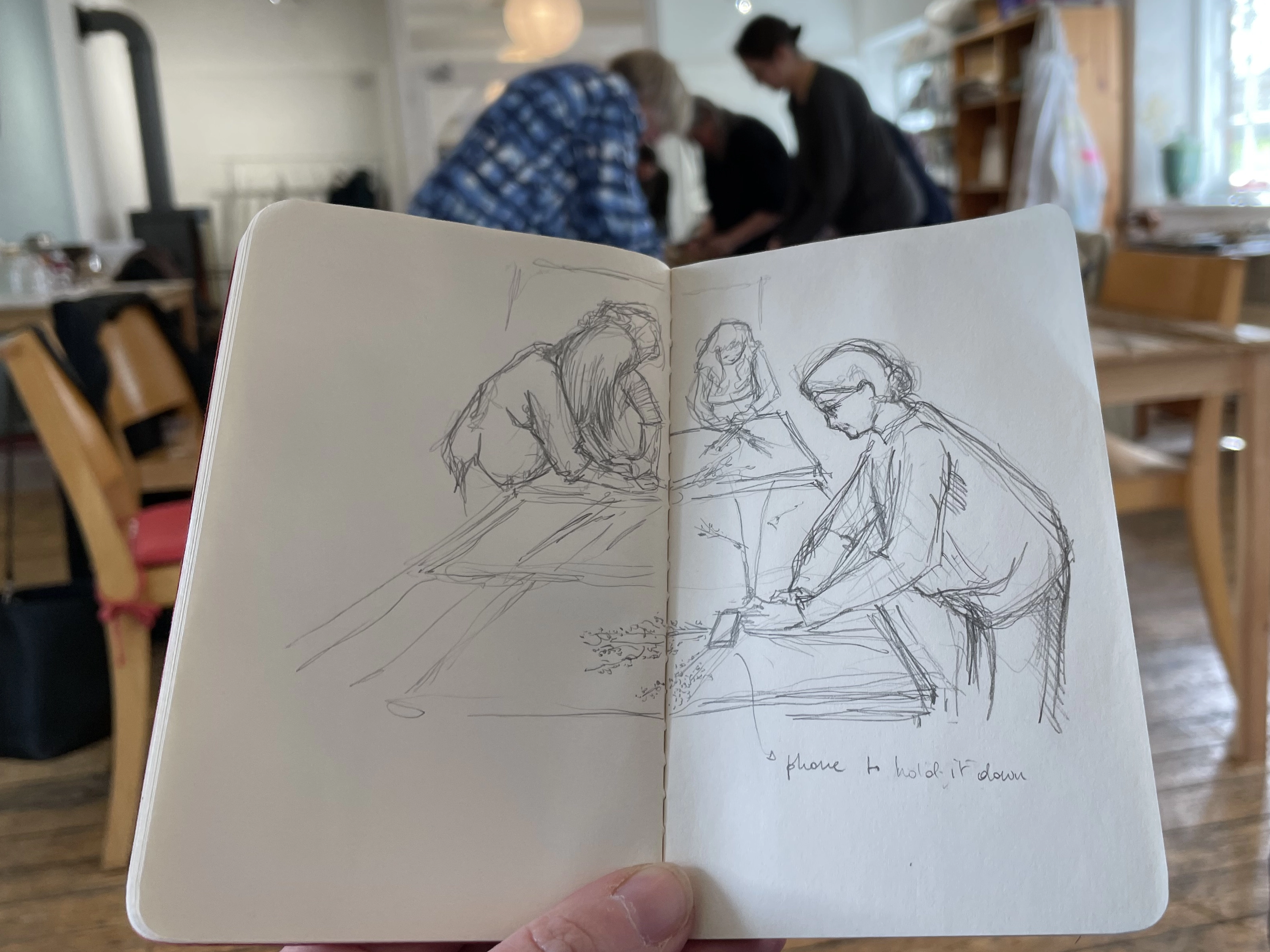 Hand holding an open sketchbook with a pencil sketch of a group of people around tables weaving flax, which can be seen blurred in the background behind the sketchbook 