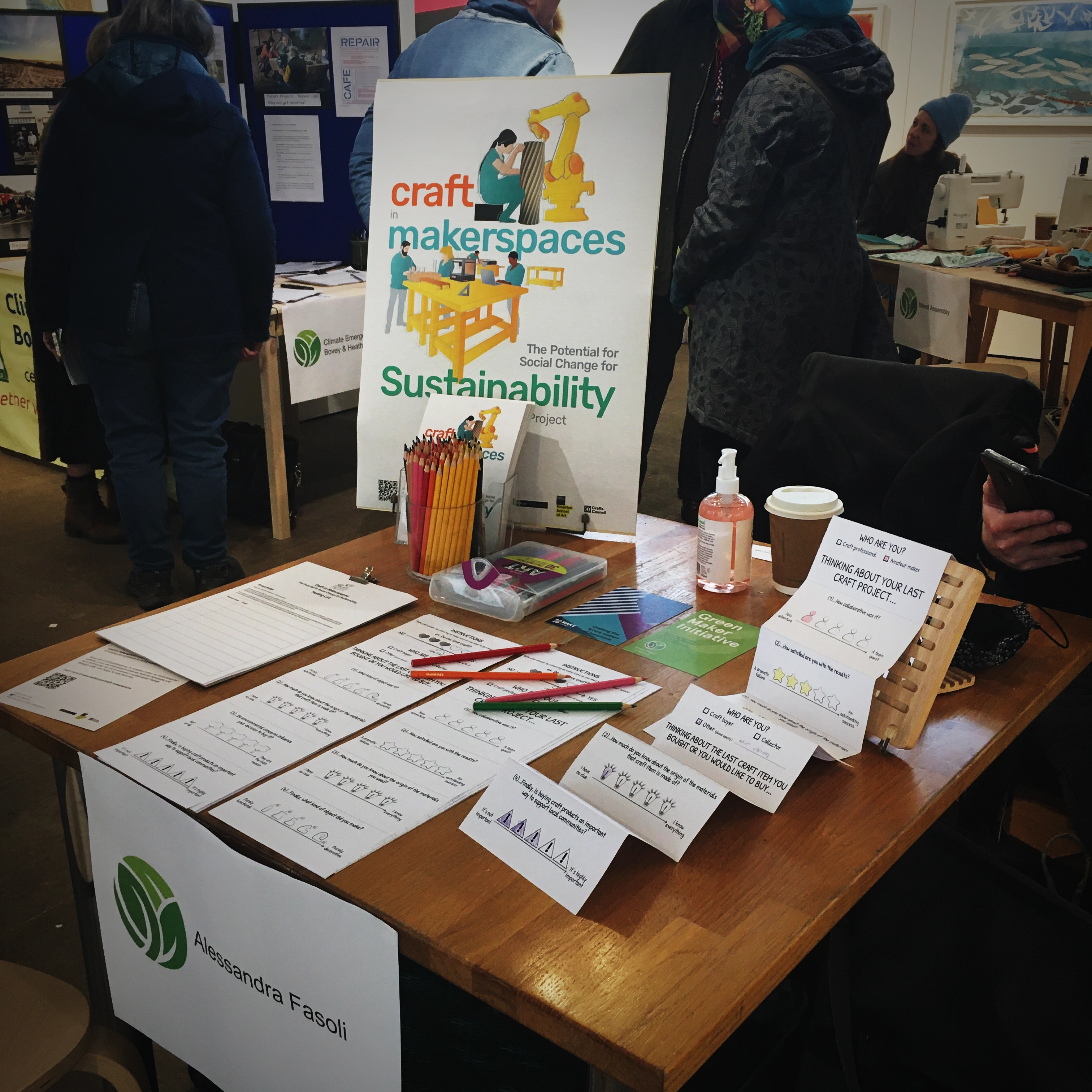 Picture of a table with concertina paper sheet of the surveys question, coloured pencils, and a poster reading 'Craft in Makerspaces. The Potential for Social Change for Sustainability'. A label on the front of the table reads 'Alessandra Fasoli' with the Green Maker Initiative logo. Indistinct people in the background 