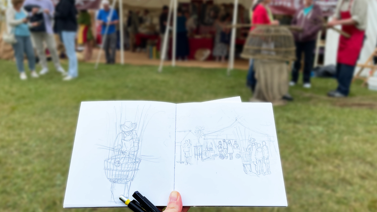 Self picture of my hand holding the sketchbook with pavillions and people in the background. The sketchbook is open showing pencil drawings of the scene