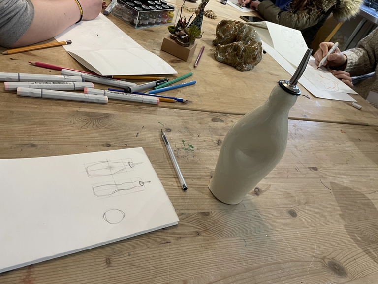 Picture of a table showing a paper with pencil drawings of a white ceramic oil dispenser placed nearby the page