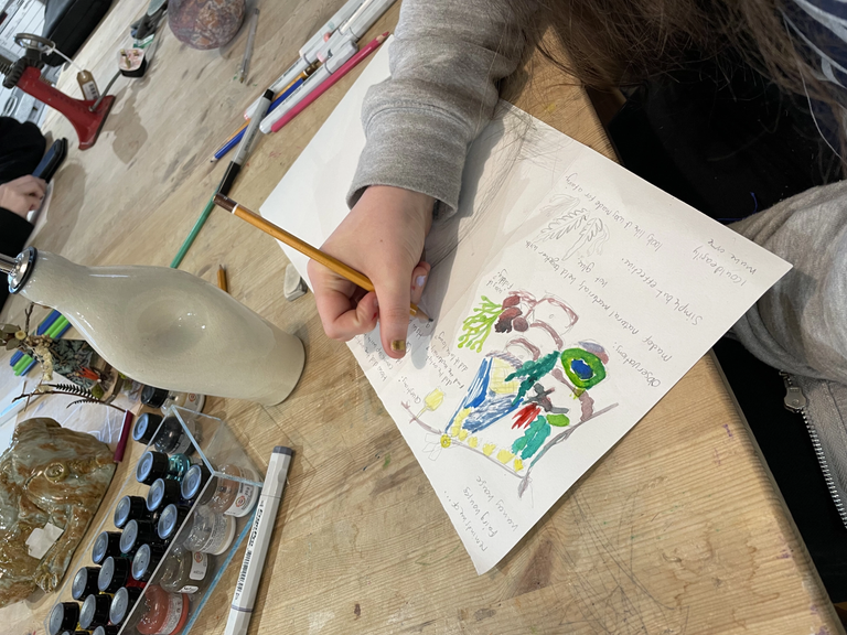 Hand holding a pencil writing down notes around a coloured ink drawing with a line of ink bottles nearby