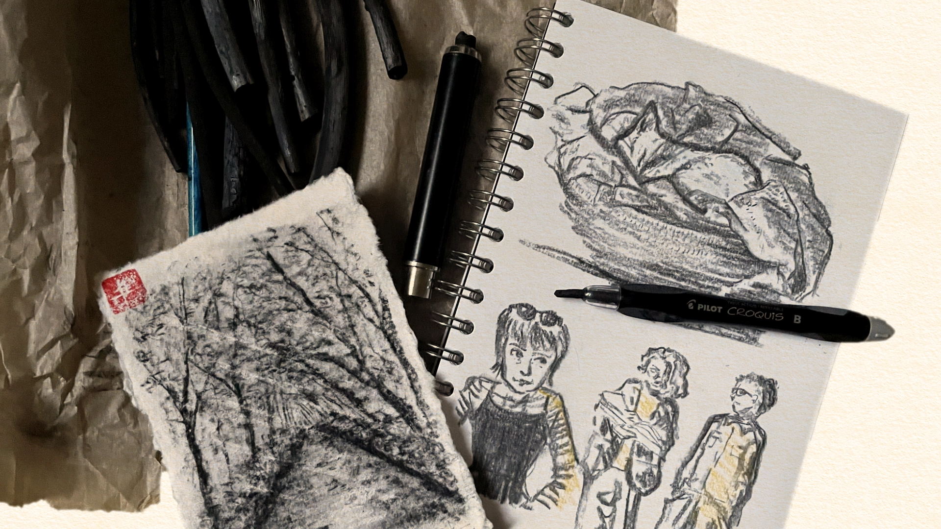 Top picture of two charcoal sketches, one on a loose white paper representing a wood path and another in a spiral sketchbook representing human figures and a cat sleeping in a round basket. They lie on a wooden floor near a batch of charcoal sticks and two clutch pencils
